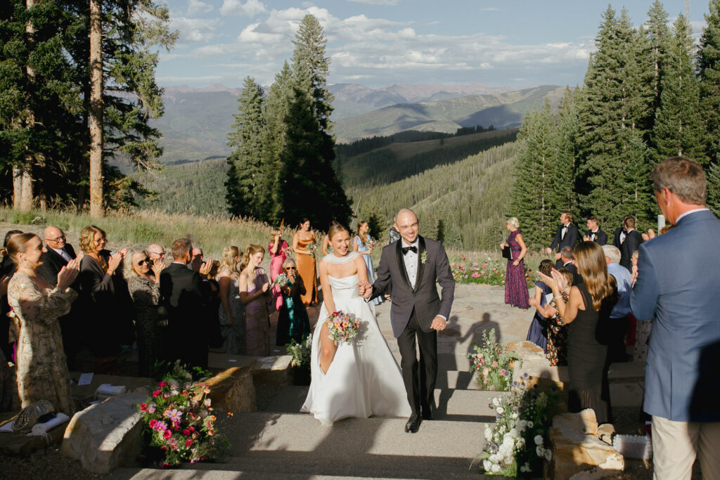 A couple walks up the steps of the  amphitheater after being married at Beaver Creek Resort, with mountains in the background and guests surrounding.
